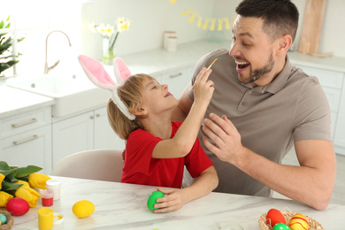 Photo of Happy son with bunny ears headband and his father having fun while painting Easter egg at table in kitchen