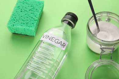 Photo of Eco friendly natural cleaners. Vinegar in bottle, sponge and jar of soda on green background