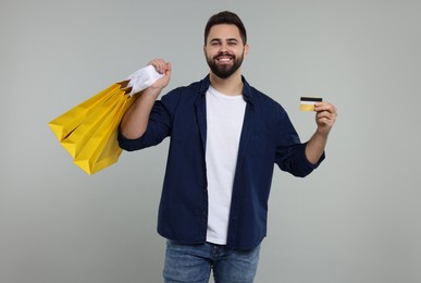 Happy man with many paper shopping bags showing credit card on grey background
