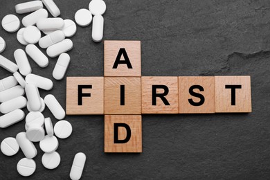 Photo of Words First Aid made of wooden cubes and pills on black table, flat lay
