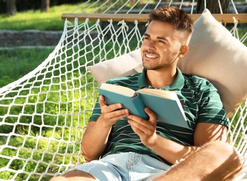 Photo of Young man reading book in comfortable hammock at green garden