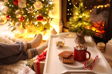 Woman sitting near fireplace at home, focus on tray with cocoa and snacks. Cozy winter holidays atmosphere