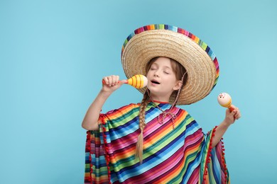 Photo of Cute girl in Mexican sombrero hat and poncho singing with maracas on light blue background