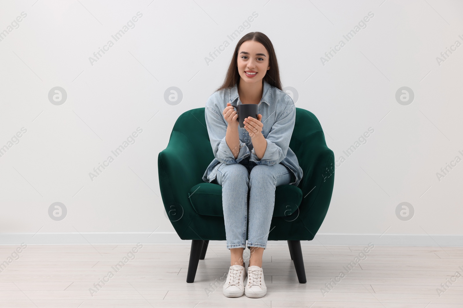 Photo of Beautiful woman with cup of drink sitting in armchair near white wall indoors