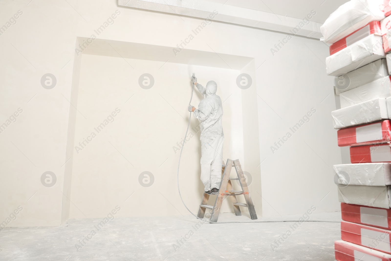 Photo of Decorator in protective overalls painting wall with spray gun indoors