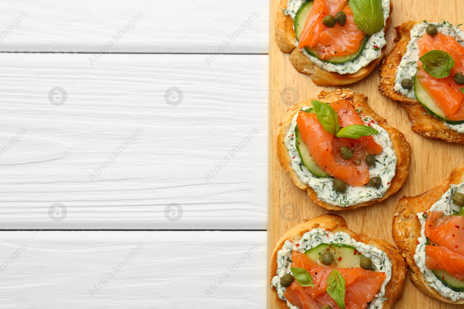 Photo of Tasty canapes with salmon, capers, cucumber and sauce on white wooden table, top view. Space for text