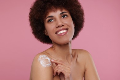 Beautiful young woman applying body cream onto shoulder on pink background