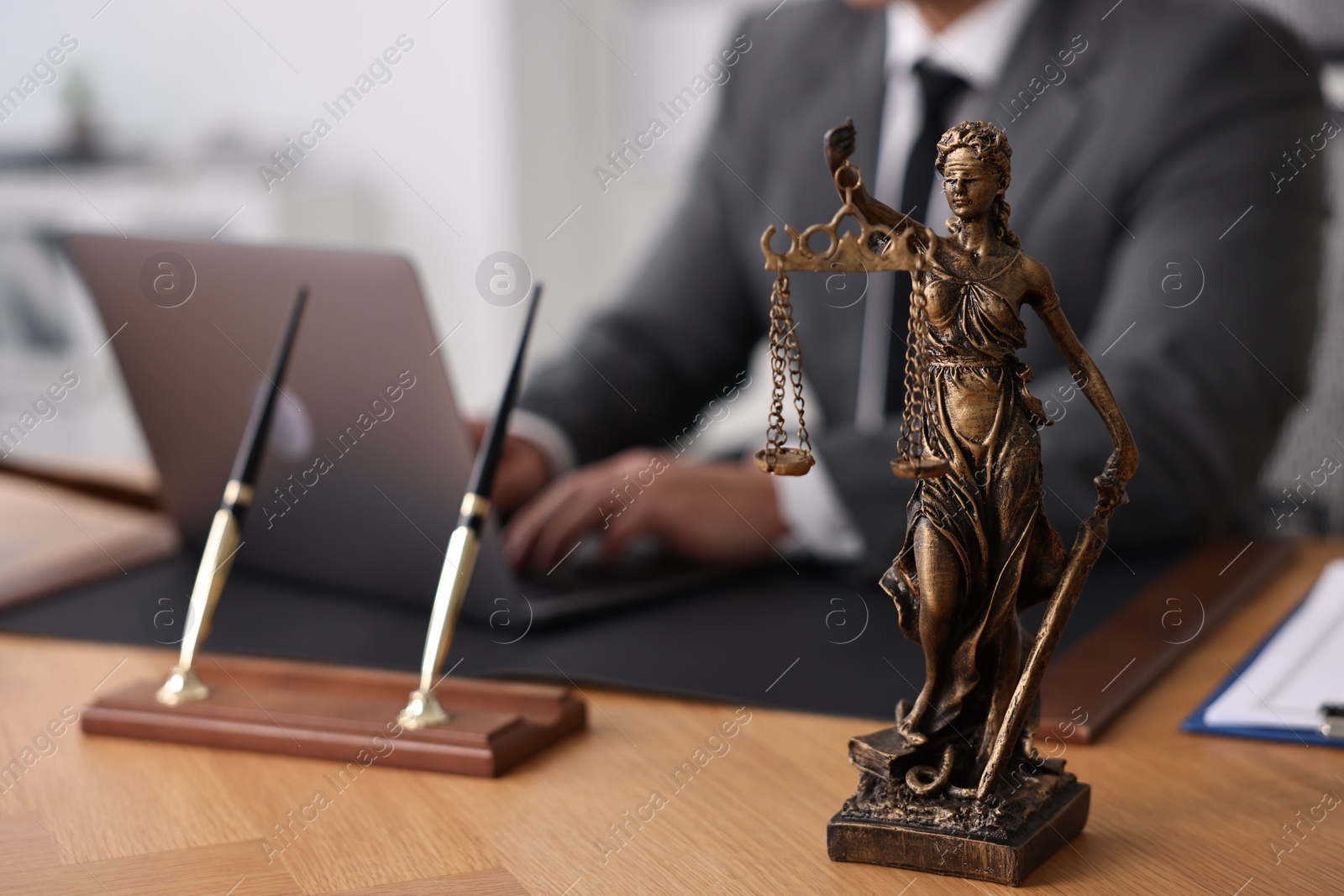 Photo of Notary working with laptop at wooden table in office, focus on statue of Lady Justice