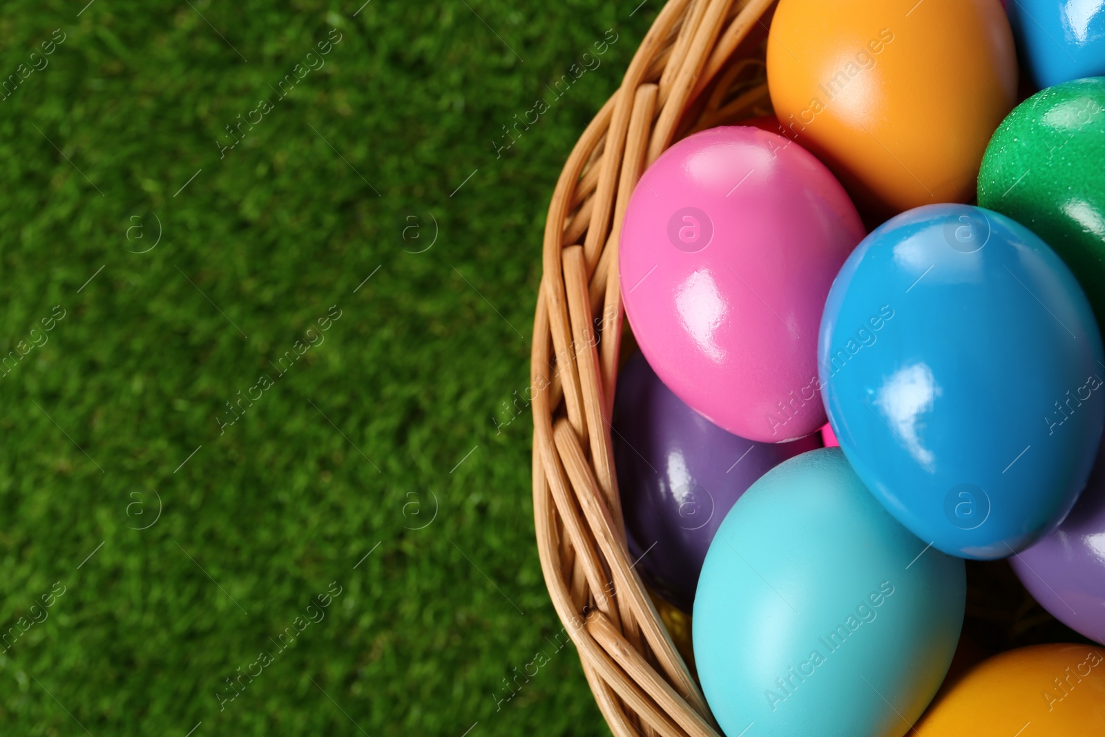 Photo of Wicker basket with bright painted Easter eggs on green grass, top view. Space for text