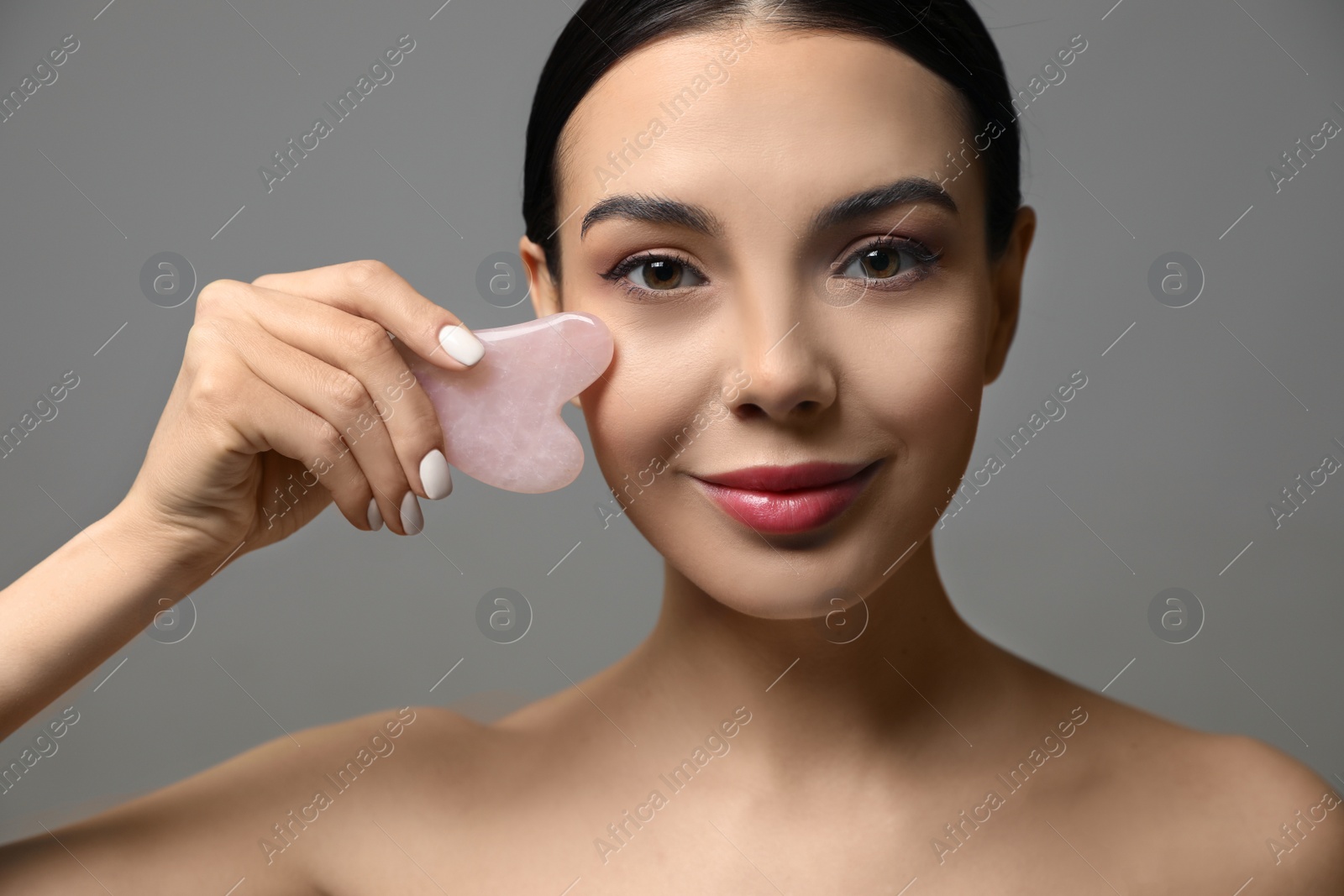 Photo of Beautiful young woman doing facial massage with gua sha tool on grey background, closeup