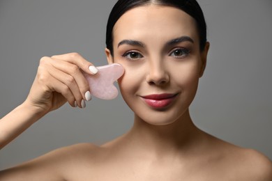 Beautiful young woman doing facial massage with gua sha tool on grey background, closeup