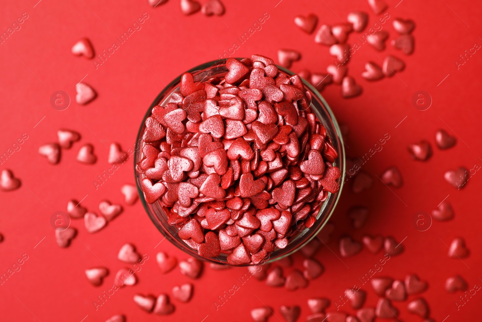 Photo of Bright heart shaped sprinkles in glass bowl on red background, flat lay