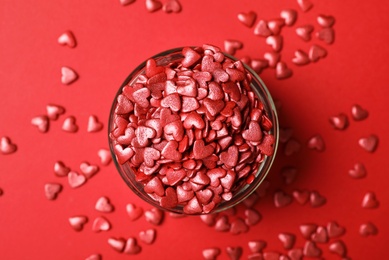 Bright heart shaped sprinkles in glass bowl on red background, flat lay