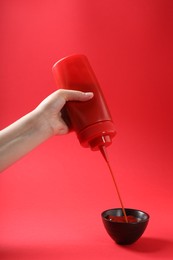 Woman pouring tasty ketchup from bottle into bowl on red background, closeup