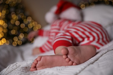 Photo of Baby in Christmas pajamas and Santa hat sleeping on bed indoors, focus on feet