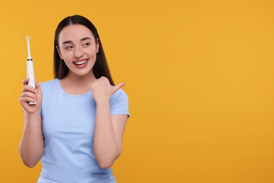 Photo of Happy young woman holding electric toothbrush and pointing on yellow background, space for text