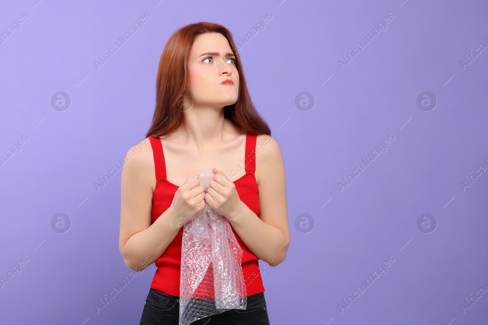 Photo of Angry woman popping bubble wrap on purple background, space for text. Stress relief