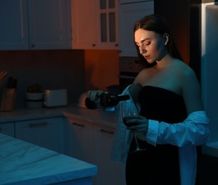 Beautiful woman pouring wine from bottle into glass in kitchen at night