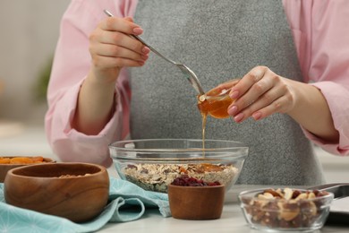 Making granola. Woman adding honey into bowl with mixture of oat flakes and other ingredients at table in kitchen, closeup