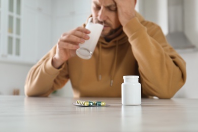 Photo of Man taking medicine for hangover at table in kitchen, focus on bottle with pills