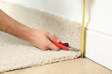 Photo of Worker with cutter knife and measuring tape installing new carpet indoors, closeup