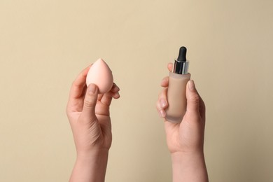 Photo of Woman holding bottle with skin foundation and makeup sponge on beige background, closeup