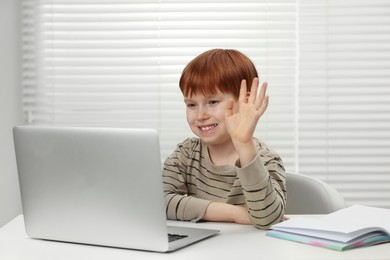 Cute boy waving hello during online lesson via laptop at white table indoors