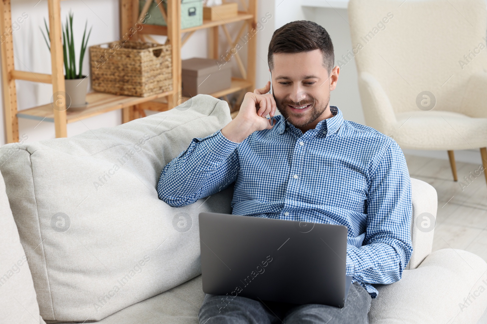 Photo of Happy man talking on phone while working with laptop on sofa at home