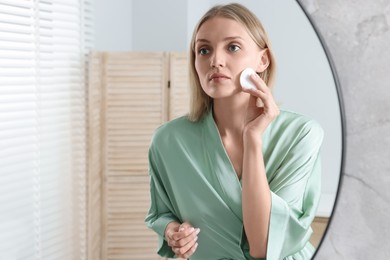 Young woman cleaning her face with cotton pad near mirror in bathroom