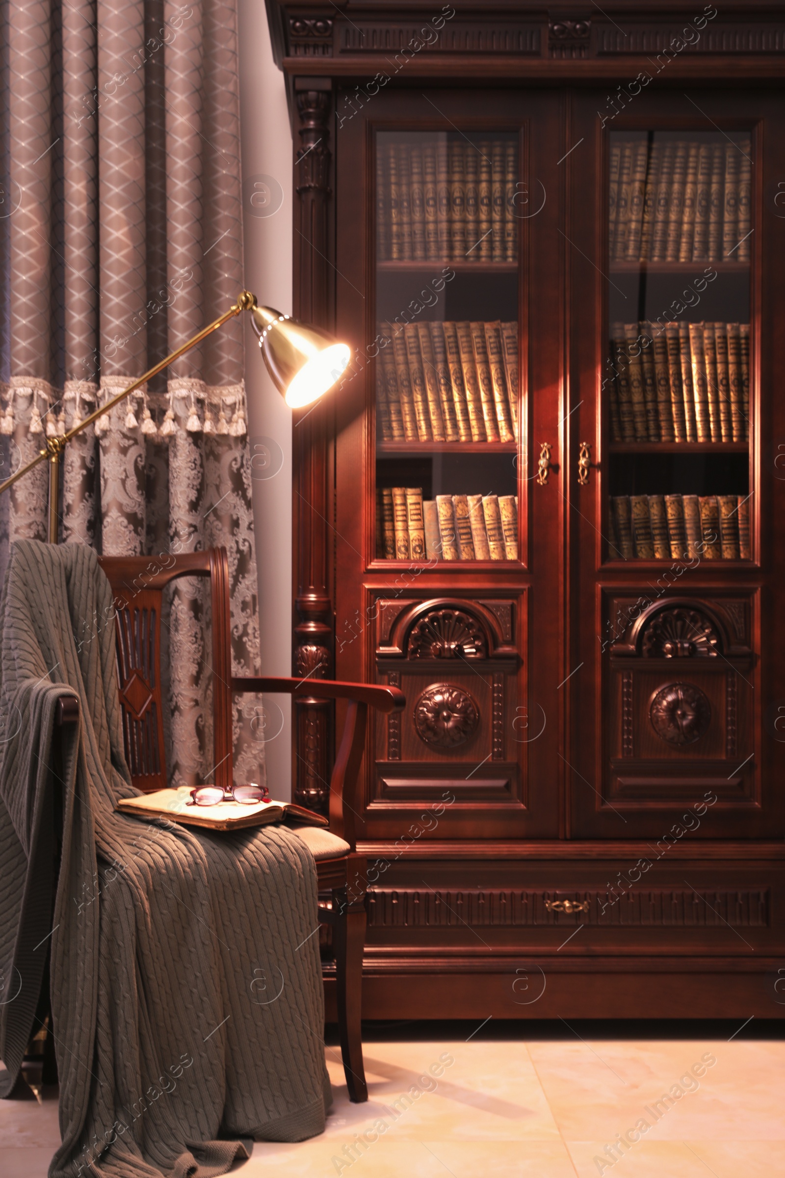 Photo of Comfortable armchair with book, blanket and lamp near wooden bookcase in library