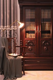 Photo of Comfortable armchair with book, blanket and lamp near wooden bookcase in library
