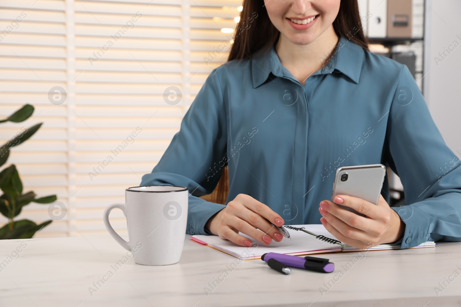 Photo of Woman taking notes while using smartphone at white marble table indoors, closeup. Space for text