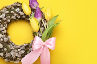 Wreath made of beautiful willow, colorful tulip flowers and pink bow on yellow background, top view. Space for text