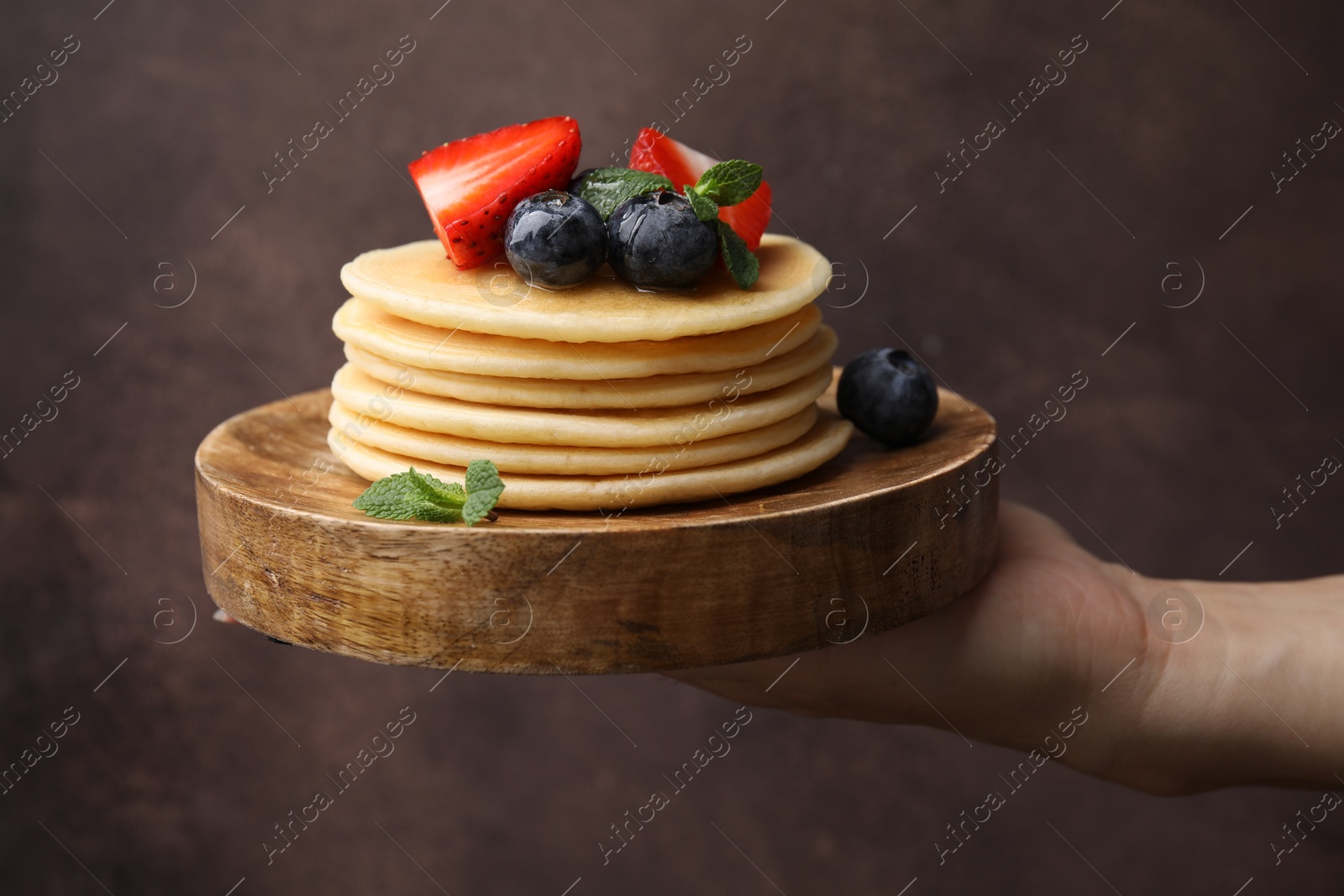 Photo of Woman holding delicious pancakes with strawberries, blueberries and mint against brown background, closeup