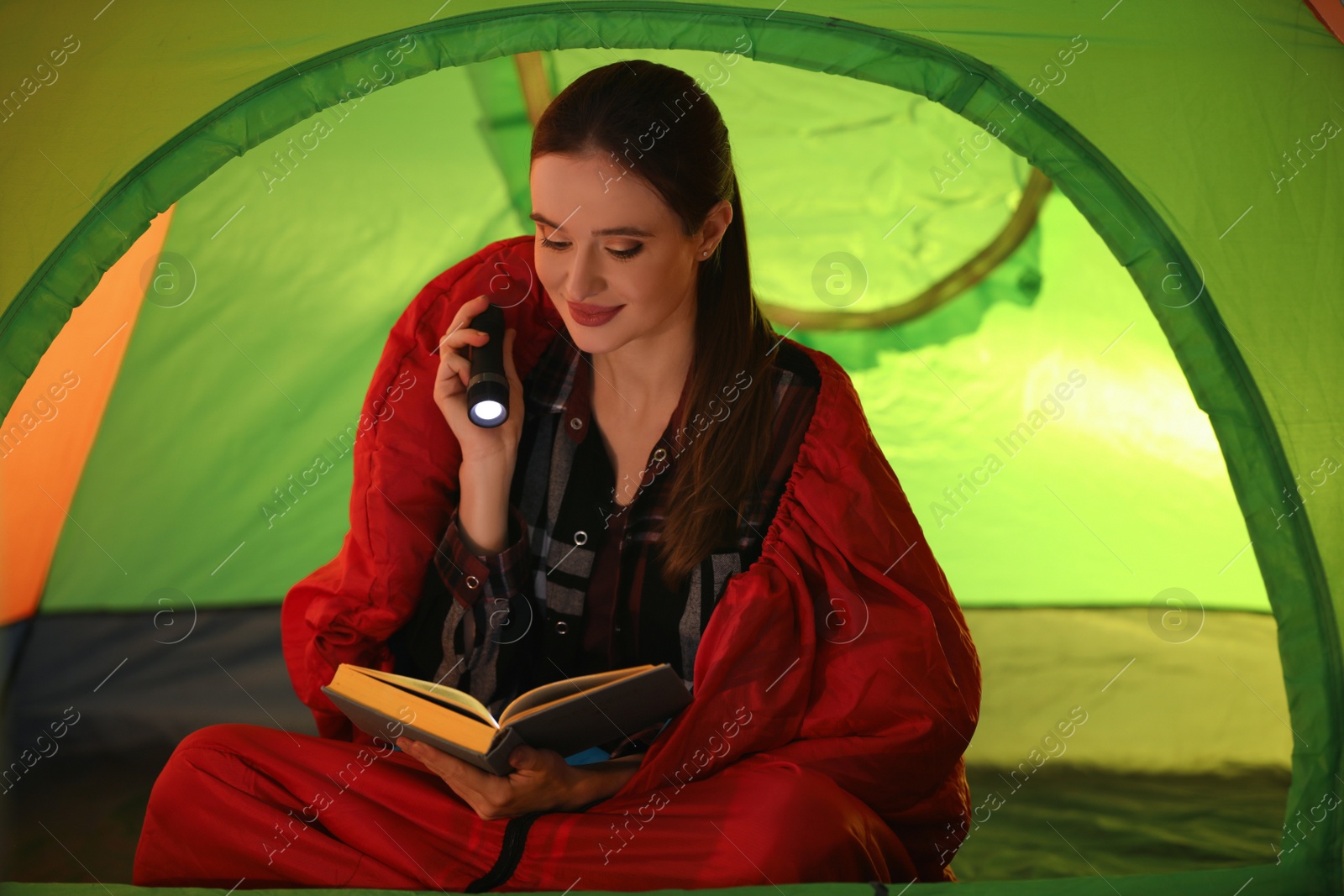 Photo of Young woman with flashlight reading book in tent