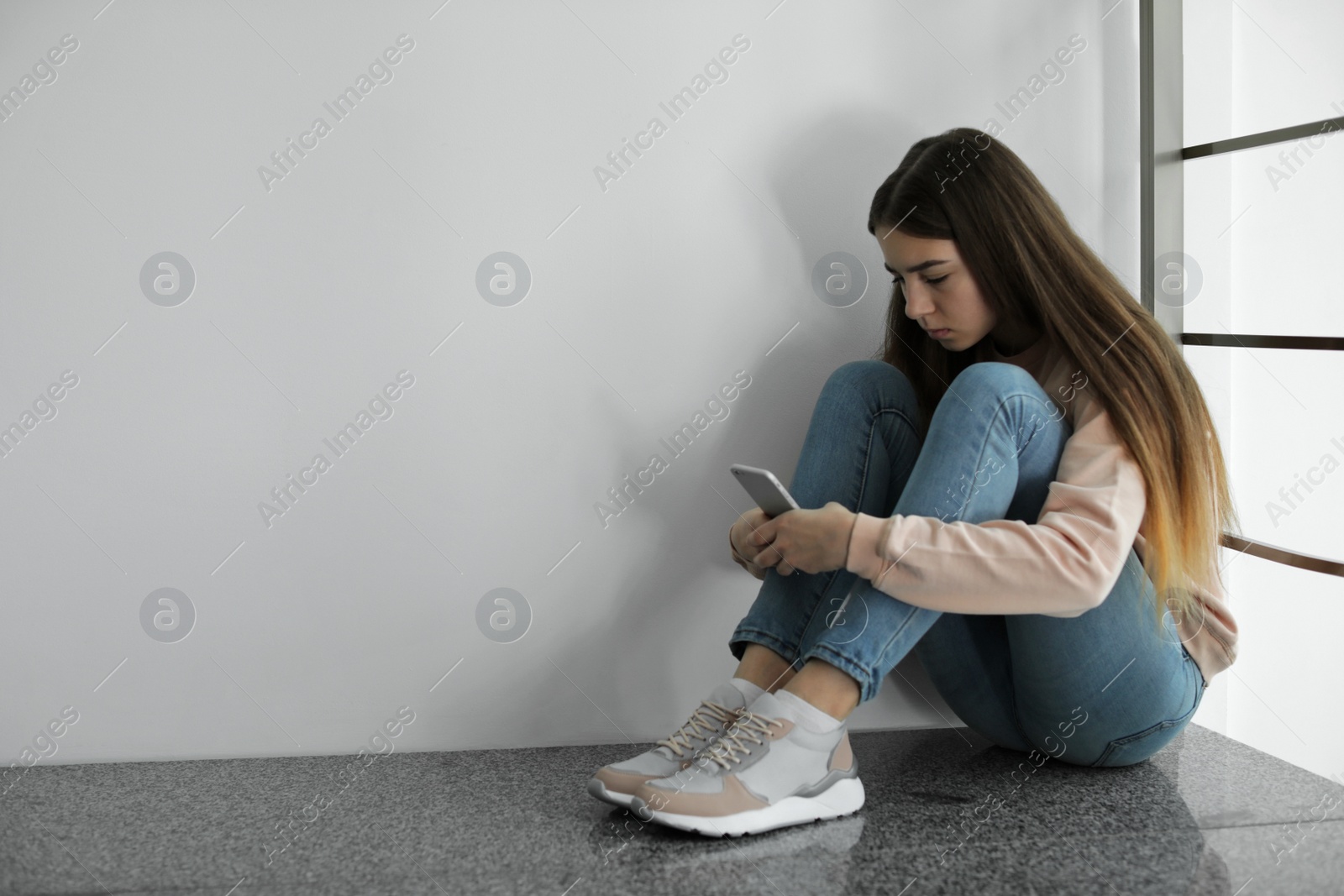 Photo of Upset teenage girl with smartphone sitting on floor near wall. Space for text
