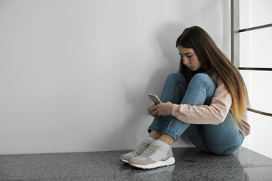 Upset teenage girl with smartphone sitting on floor near wall. Space for text