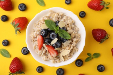 Photo of Tasty oatmeal with strawberries, blueberries and almond petals in bowl surrounded by fresh berries on yellow background, flat lay