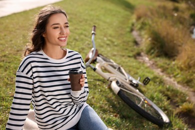 Photo of Young woman sitting on green grass and holding cupcoffee near bicycle outdoors, space for text