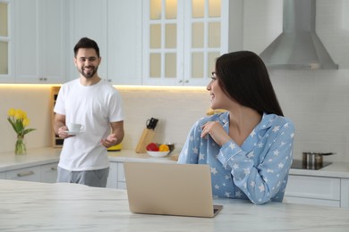 Photo of Young woman in pajamas using laptop while her boyfriend bringing coffee at home