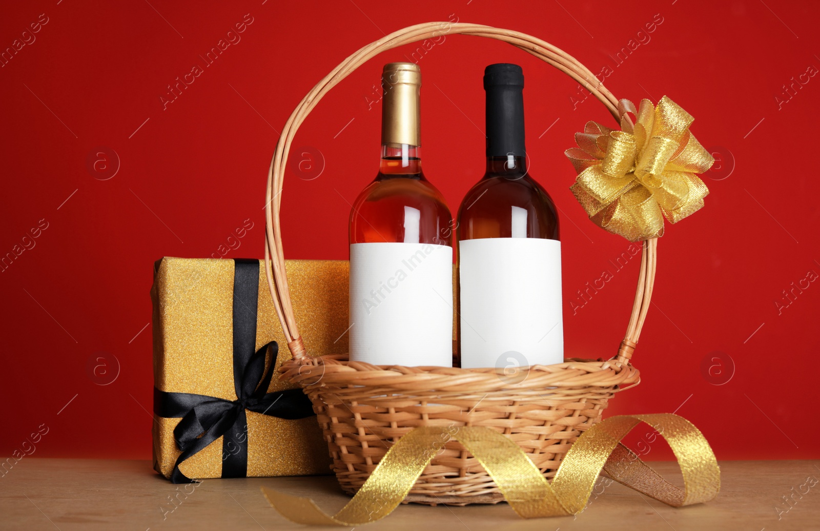 Photo of Bottles of wine in wicker basket with bow and gift on table against color background