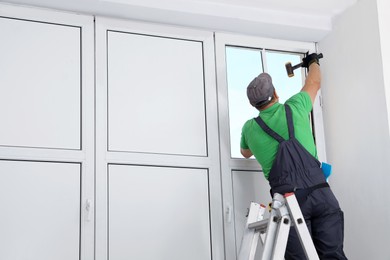 Worker on folding ladder installing window indoors, back view