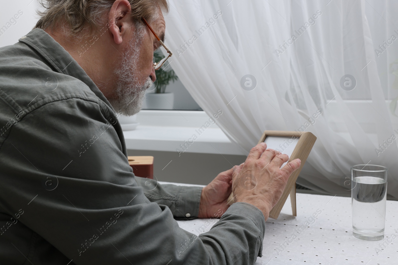 Photo of Upset senior man with photo frame at table in room. Loneliness concept