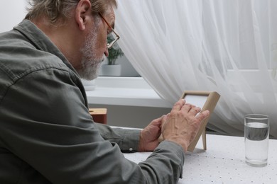 Upset senior man with photo frame at table in room. Loneliness concept