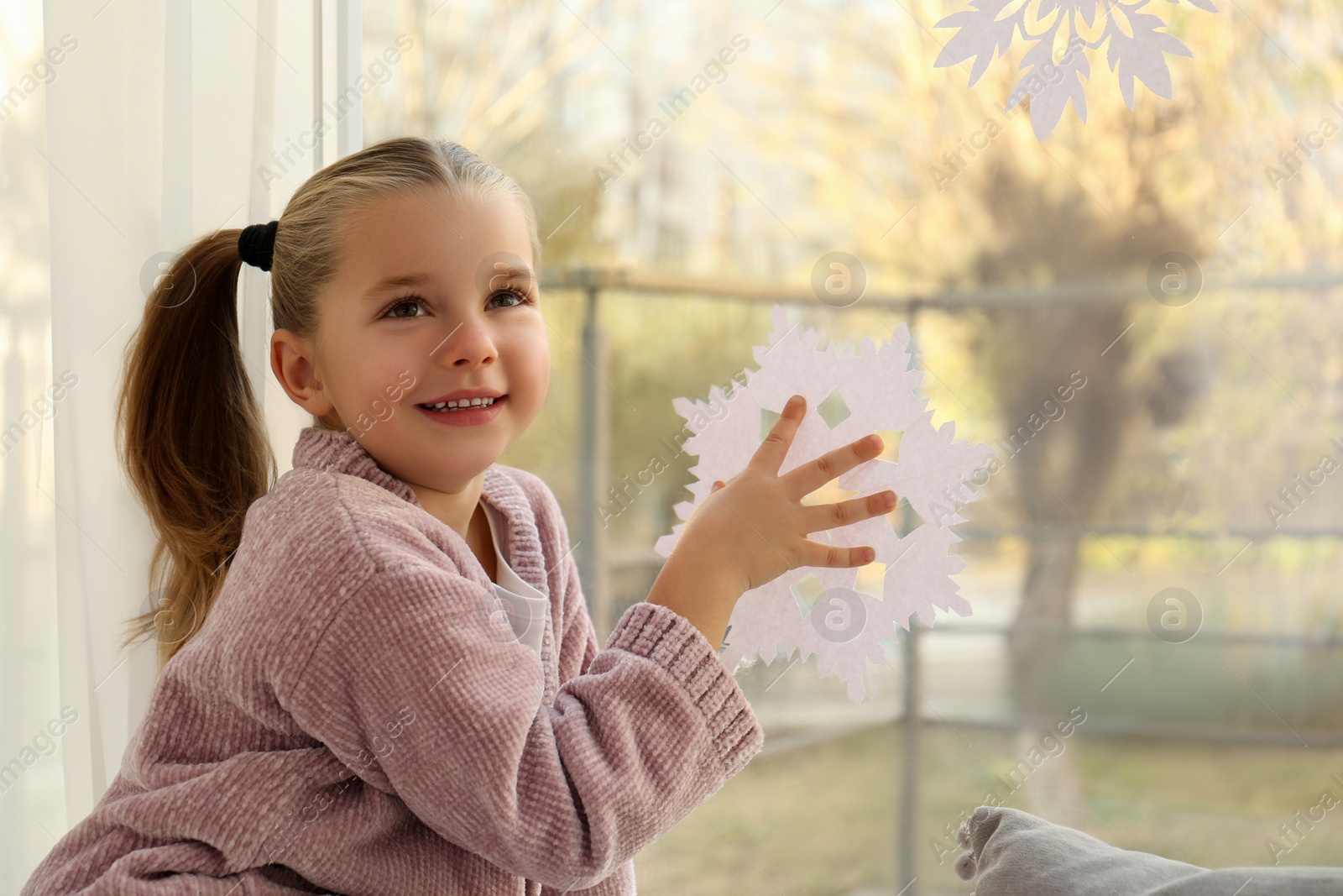 Photo of Little girl decorating window with paper snowflake indoors