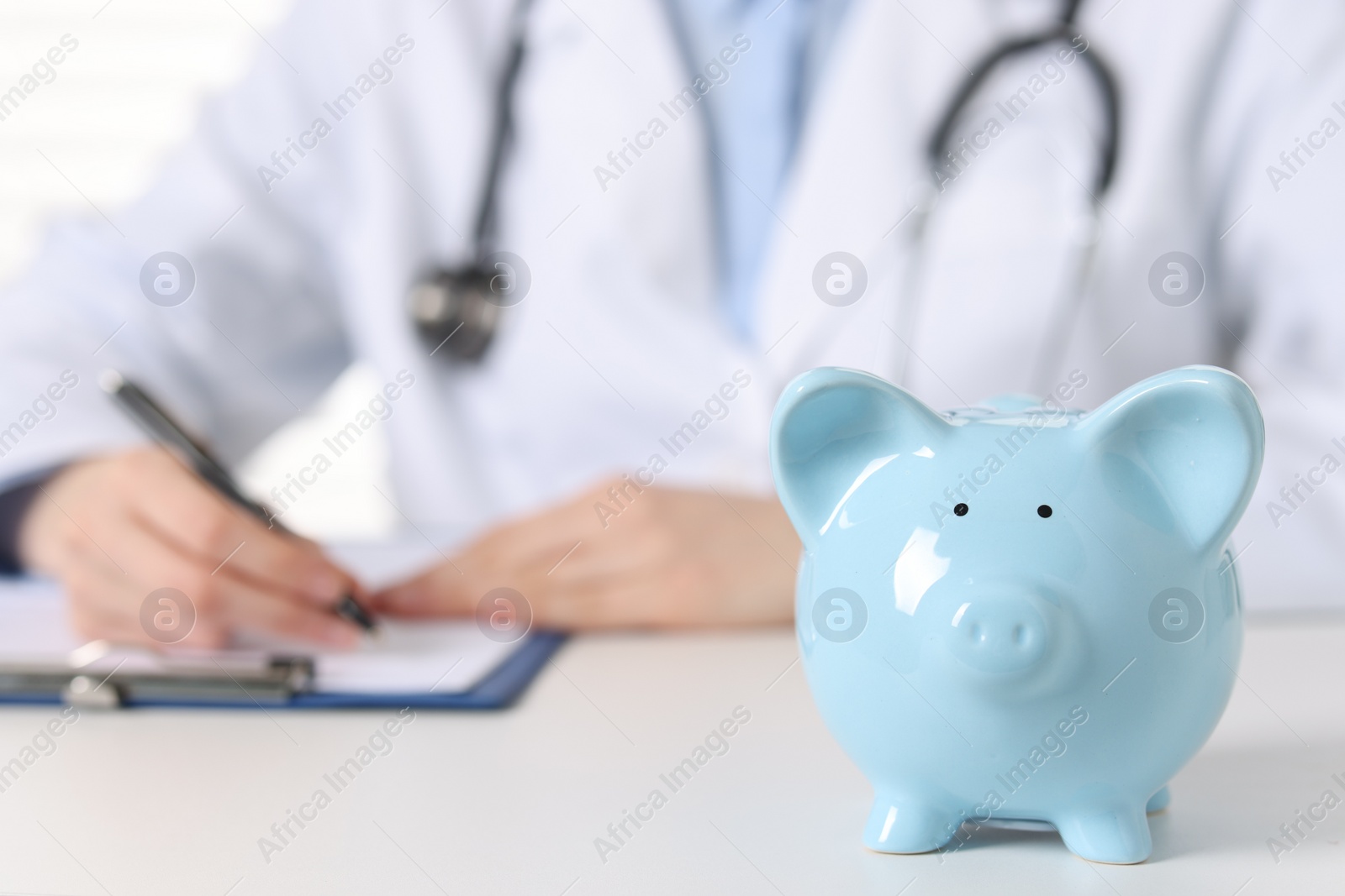 Photo of Doctor making notes at white table, focus on piggy bank