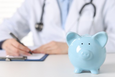 Doctor making notes at white table, focus on piggy bank