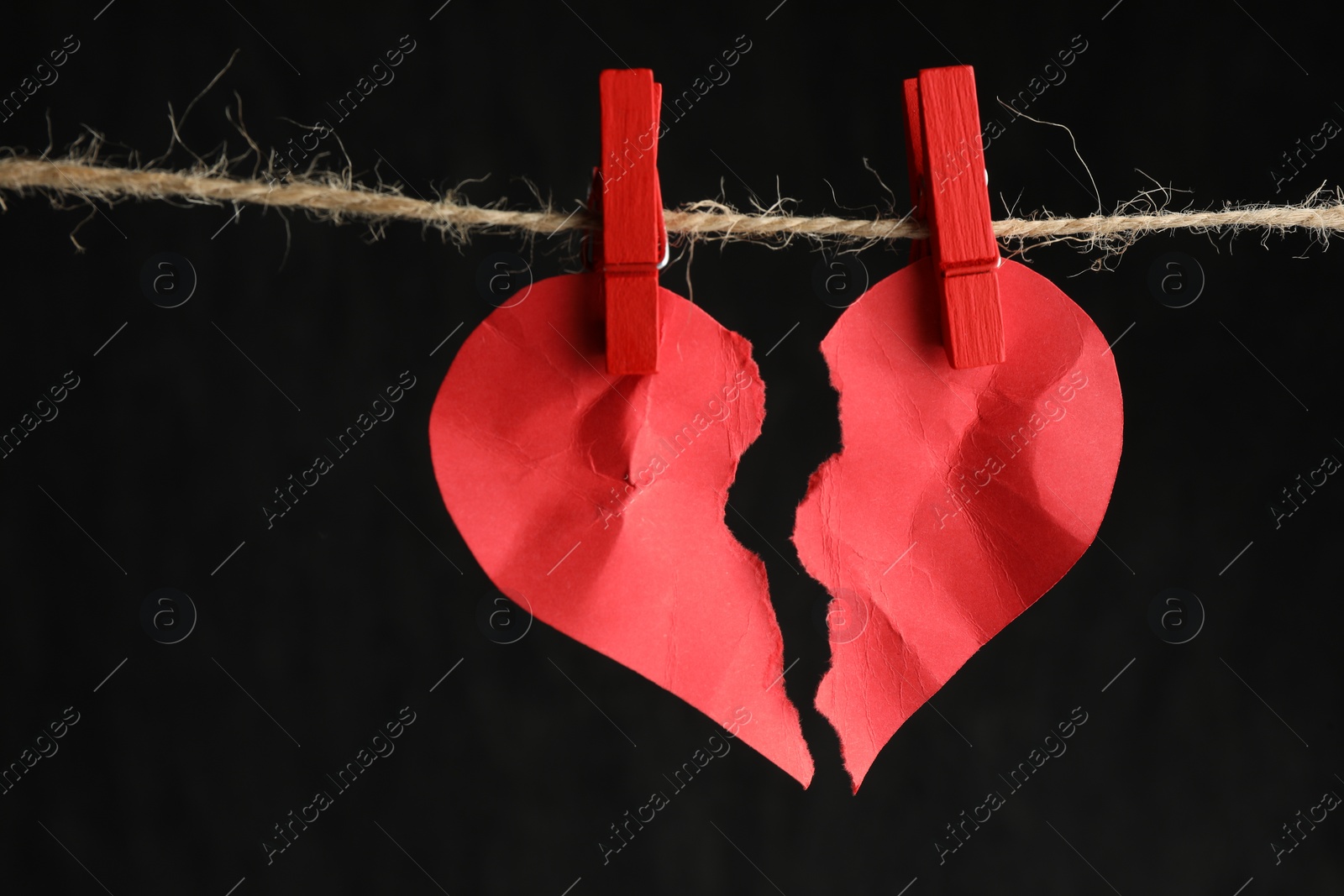 Photo of Halves of torn red paper heart on rope against black background, closeup. Broken heart