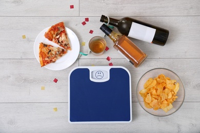 Photo of Digital scales, junk food and alcohol on wooden floor after party, top view