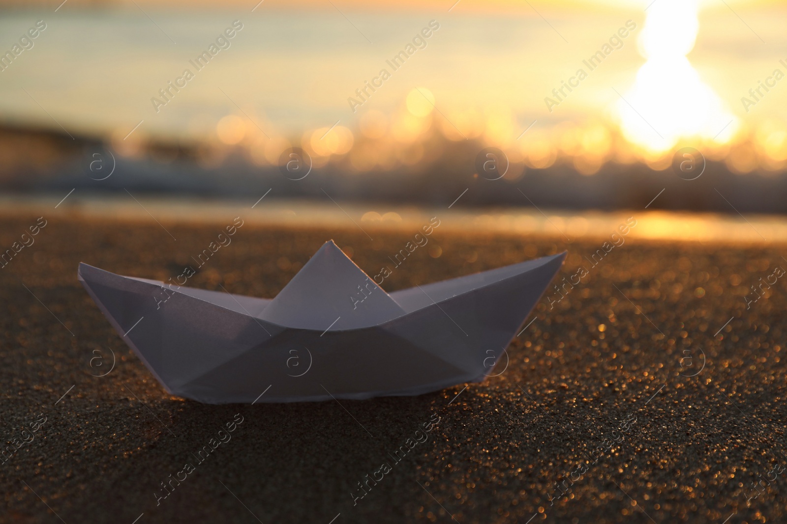 Photo of White paper boat on sand near sea at sunset, closeup. Space for text